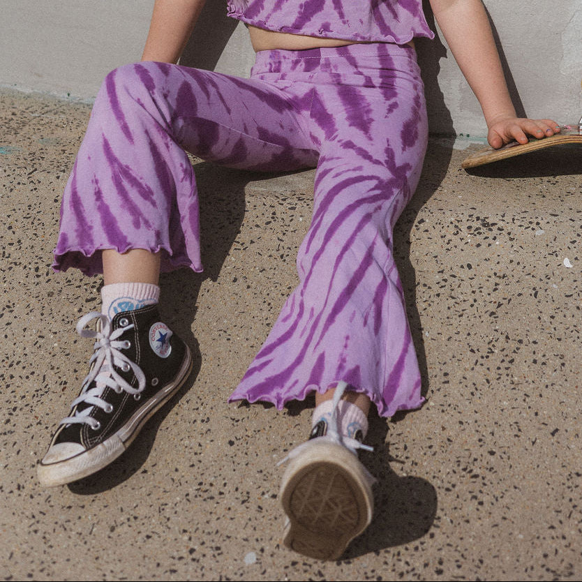 Child wearing a purple tie-dye outfit sitting on the ground against a plain wall.