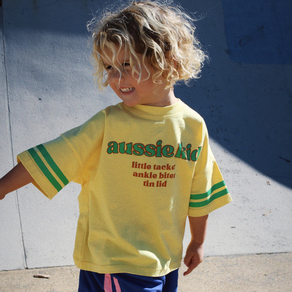 Child wearing a yellow 'aussie kid' t-shirt and blue shorts on a concrete surface.