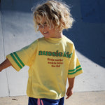 Child wearing a yellow 'aussie kid' t-shirt and blue shorts on a concrete surface.