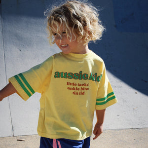 Child wearing a yellow 'aussie kid' t-shirt and blue shorts on a concrete surface.