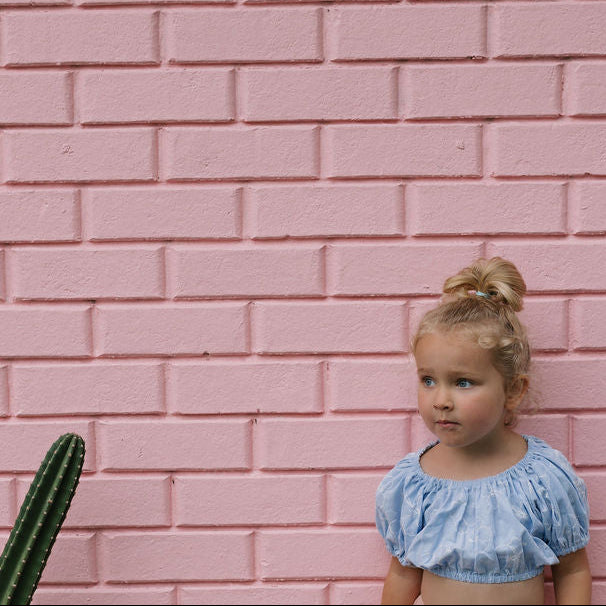 Young girl in a blue top and pink skirt standing against a pink brick wall with potted plants.