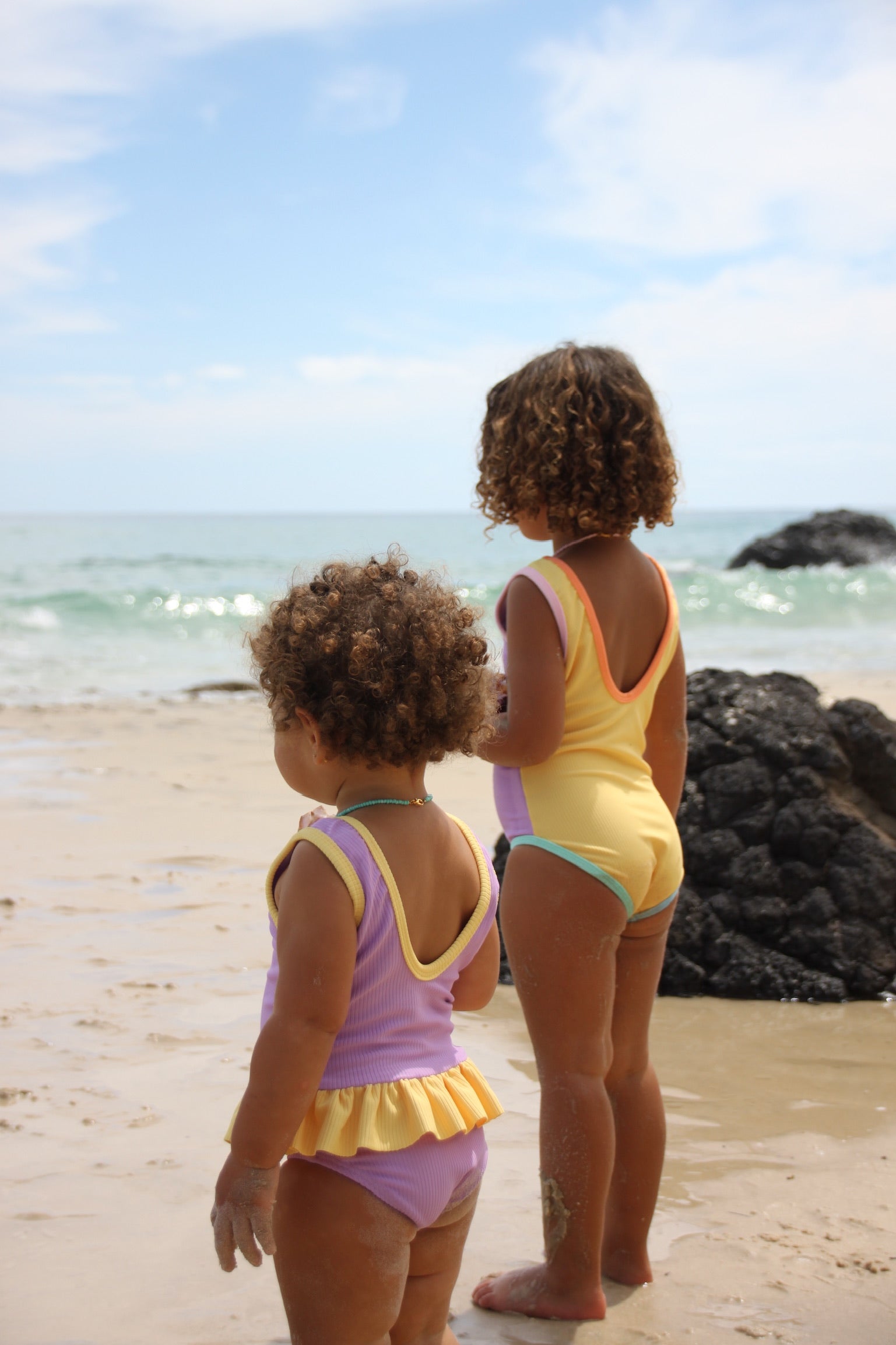 Two children in swimsuits standing on a beach looking at the ocean.