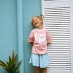 Child wearing a pink shirt with text standing against a light blue wall.