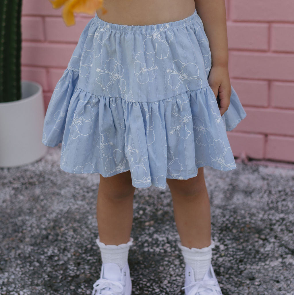 Child wearing a light blue floral skirt and white sneakers on a textured surface with a pink wall in the background.