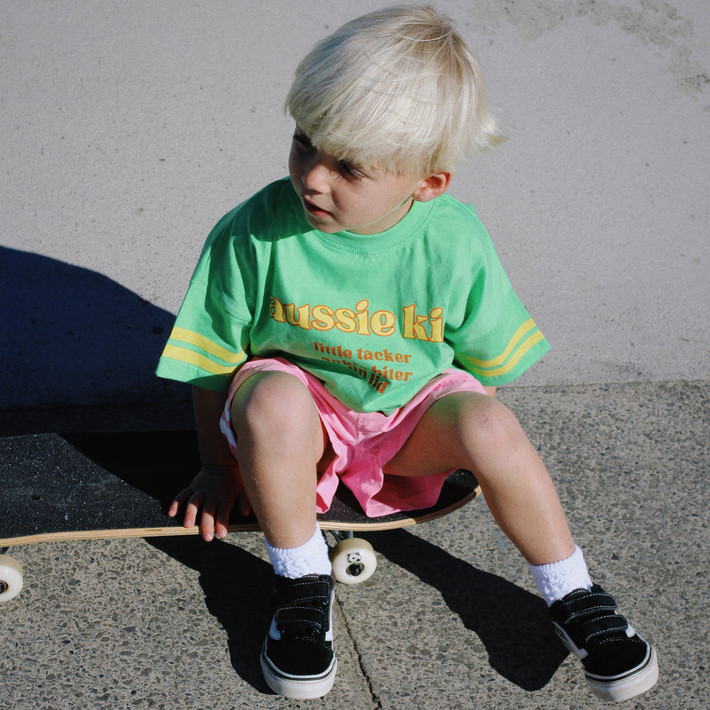 Child sitting on a skateboard wearing a green t-shirt with text and pink shorts.
