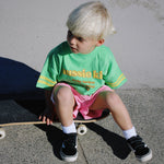 Child sitting on a skateboard wearing a green t-shirt with text and pink shorts.