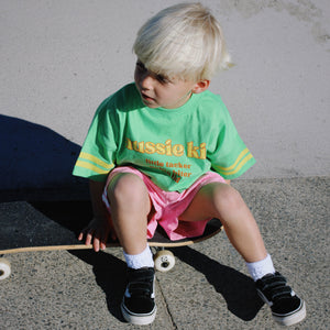 Child sitting on a skateboard wearing a green t-shirt with text and pink shorts.